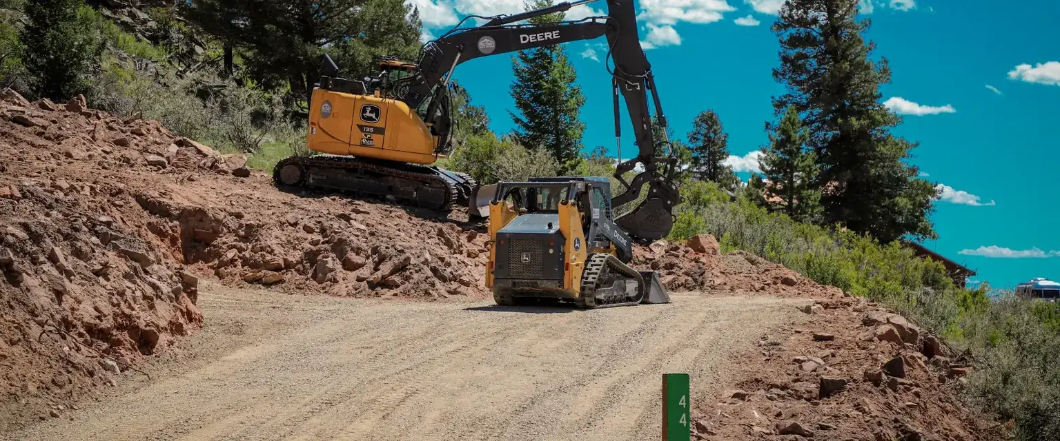 Photo: a Deere excavator and a compact track loader cutting a road through a cleared mountain lot in Park County