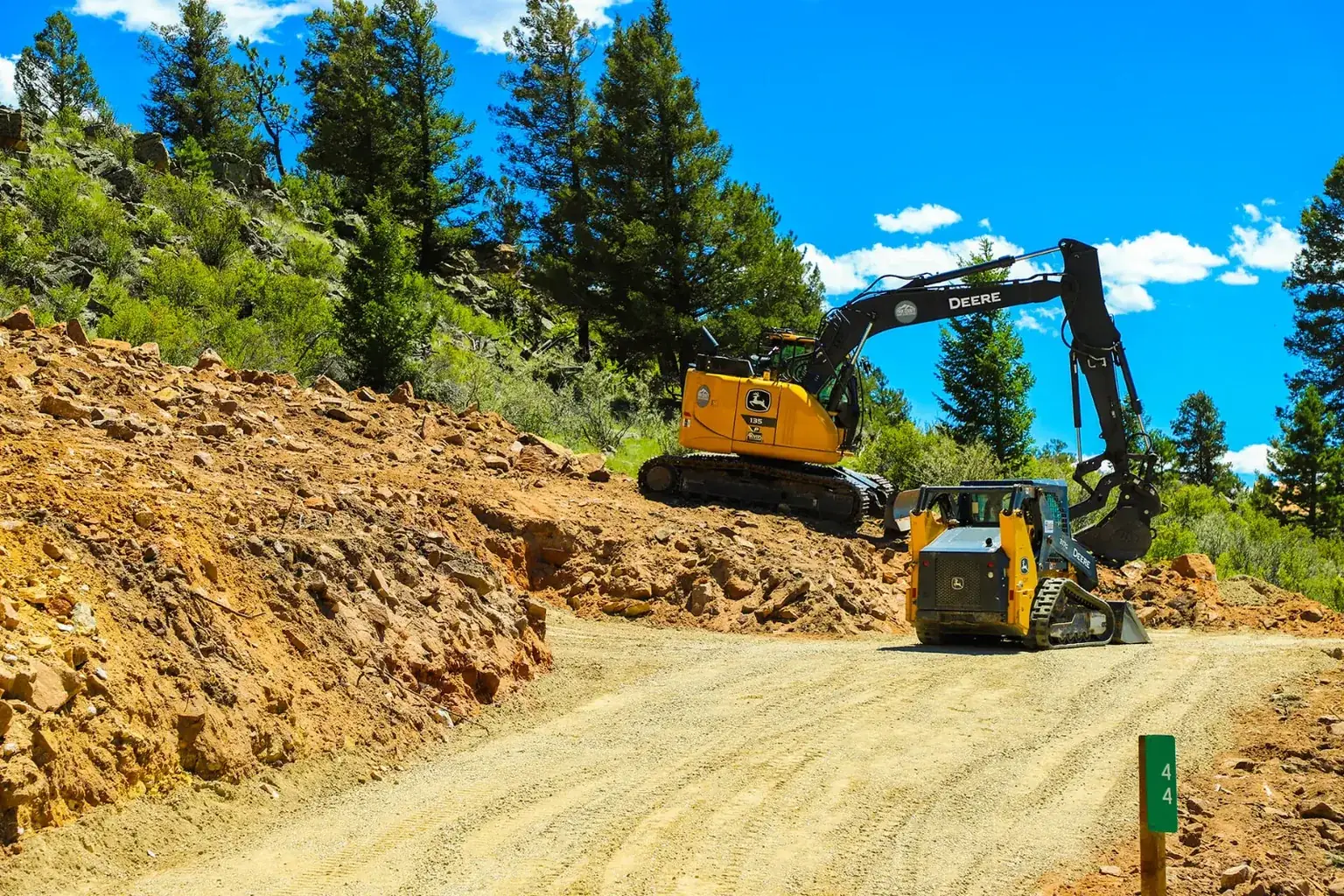 Photo: a Deere excavator and a compact track loader cutting a gravel road through a mountain lot in Park County, Colorado
