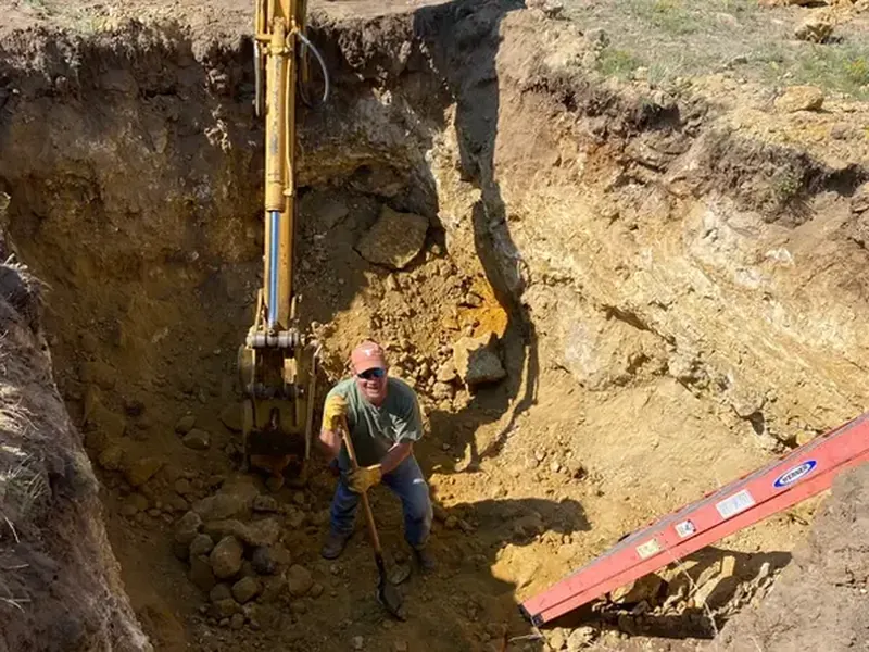 Photo: an operator standing in a deep utility trench beside an excavator bucket