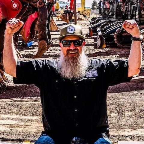 Photo: Phil George in a Park County Gravel cap and sunglasses, arms up, in front of a row of excavators